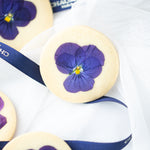 A closeup on one of the botanical shortbread cookies. The cookie is a pale, sandy colour, and it has a purple edible viola flower pressed into it for a pop of colour.