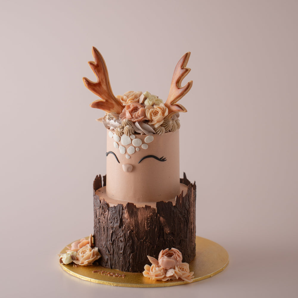A two tier cake in a rustic style. The top tier looks like a deer's head. It has hand piped buttercream flowers on top, and two cookie antlers sticking out. It has a cute face with smiling eyes. The bottom tier has chocolate wrapped around that looks like a tree log. There are buttercream flowers on the cakeboard.
