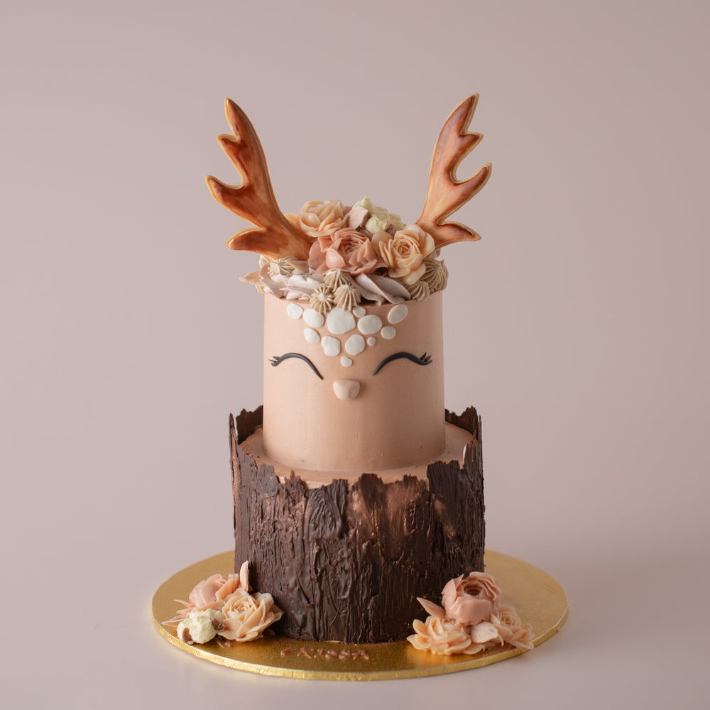A two tier cake in a rustic style. The top tier looks like a deer's head. It has hand piped buttercream flowers on top, and two cookie antlers sticking out. It has a cute face with smiling eyes. The bottom tier has chocolate wrapped around that looks like a tree log. There are buttercream flowers on the cakeboard.