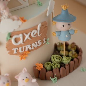 
                  
                    Load image into Gallery viewer, A close up on a fondant scarecrow standing in a cabbage and pumpkin patch. The scarecrow has a blue hat and clothes. Next to the scarecrow in the middle of the cake there is a brown fondant &amp;quot;wood&amp;quot; sign board. It says &amp;quot;Axel turns 1&amp;quot;.
                  
                
