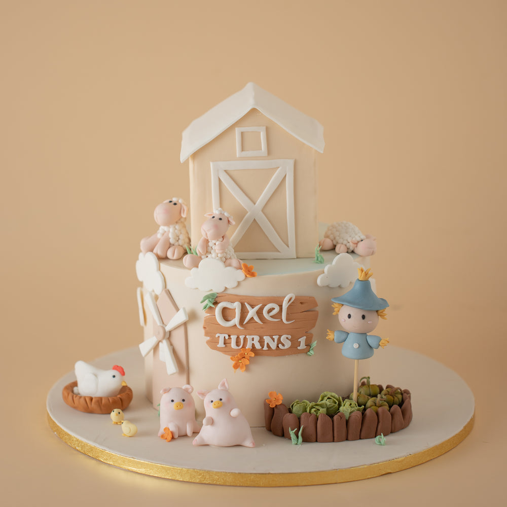 A two tier farm theme cake in neutral shades. The top tier has been carved and decorated to look like a beige and white barn. The bottom tier has a fondant windmill at the side, along with 3D fondant farm animals and a cute scarecrow. There is a fondant cabbage and pumpkin patch on the cake board.