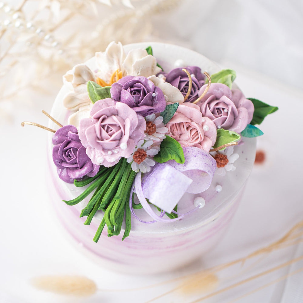 
                  
                    Load image into Gallery viewer, A white buttercream cake with streaks of purple buttercream, and various shades of purple buttercream roses on top of the cake. The buttercream roses have green stems, and resemble a bouquet of flowers. The cake has some edible sugar pearls scattered on top.
                  
                