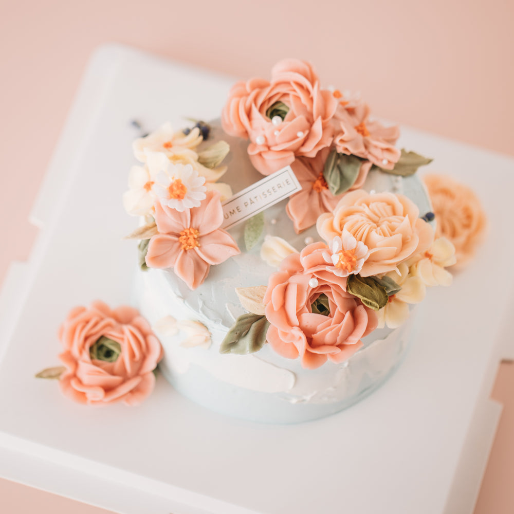 
                  
                    Load image into Gallery viewer, A light, dusty blue buttercream cake with pink and coral buttercream flowers on top, along with green edible wafer leaves. The flowers are assorted and realistic.
                  
                