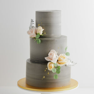 
                  
                    Load image into Gallery viewer, A three tier wedding cake with a concrete grey buttercream base. The buttercream has streaks of lighter grey marbled into it. There are some light pink and ivory edible sugar roses in the middle, along with some pale edible green sugar leaves. The flowers look incredibly realistic.
                  
                