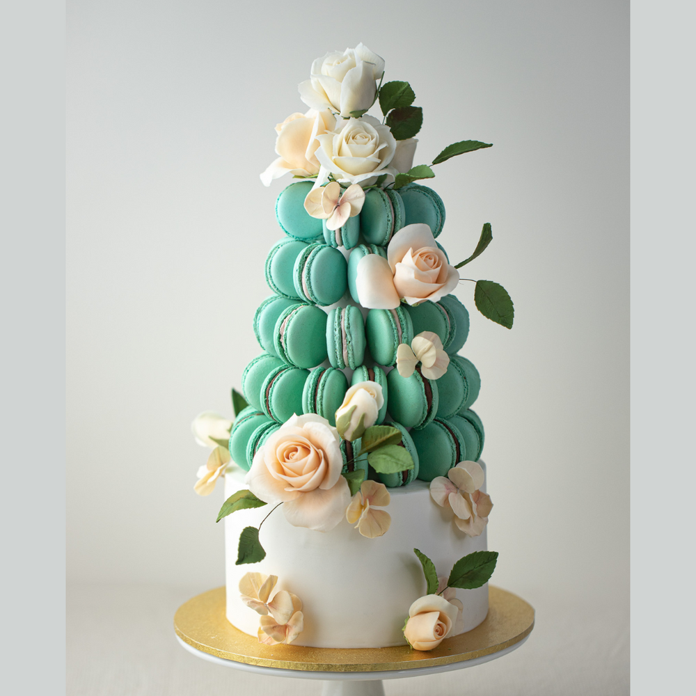 A macaron tower sitting on a white wedding cake. The macaron tower is covered in teal coloured macarons, and is adorned with numerous white and ivory edible sugar roses, along with some dark green edible sugar leaves. The cake below the macaron is a simple white buttercream cake, and also has the edible sugar roses.