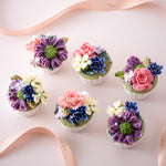 6 cupcakes with assorted hand piped buttercream flowers in vibrant pink, purple, blue and ivory. There are sugar pearls lightly sprinkled on top. The flowers look incredibly realistic and beautiful.