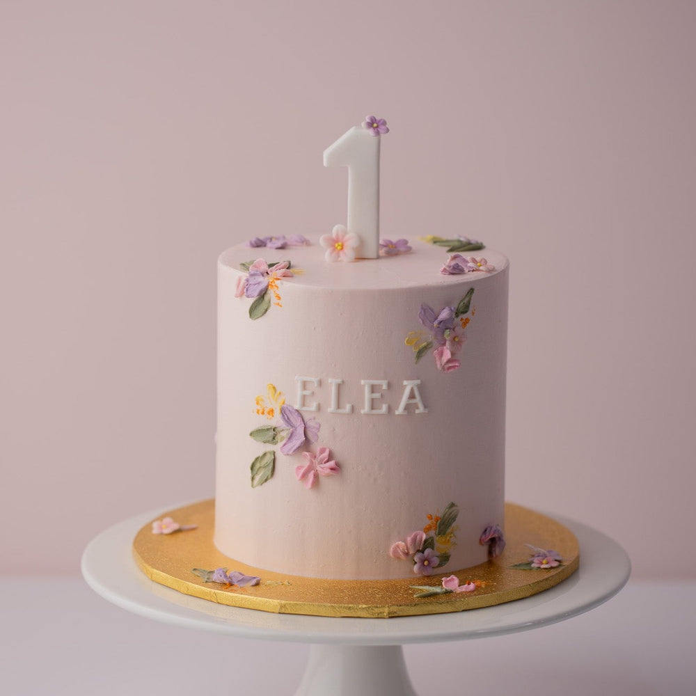 Light pink buttercream cake with little palette knife painted flowers on top and the sides of the cake. The palette knife buttercream flowers are purple, pink and yellow. There is a fondant number One on top of the cake, and the word "flea" in fondant on the front of the cake.