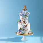 A two tier Longevity theme cake. There are two hand painted cranes on the cake, along with some hand painted branches and floral details. On the top of the cake, there are two large sugar peonies, and all around the cake there are small sugar hydrangeas. There are five small fondant peaches around the cake