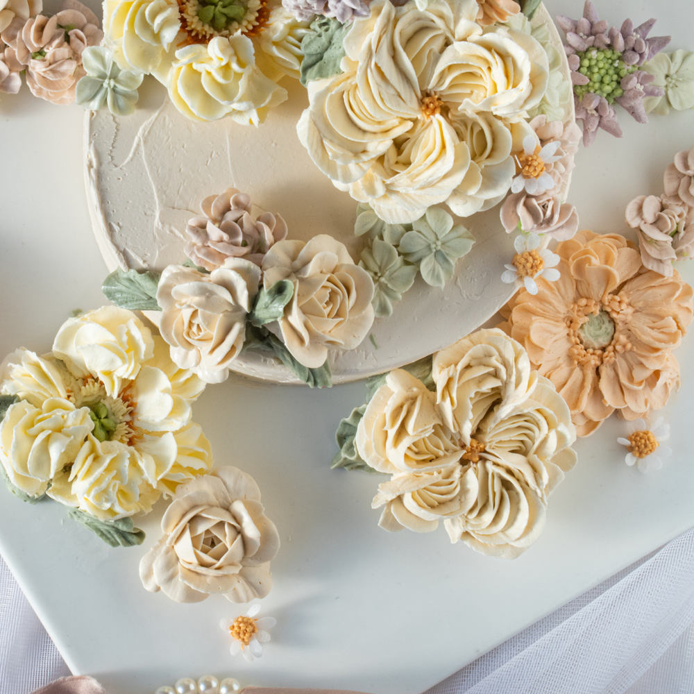 Light ivory buttercream cake with numerous buttercream flowers in white, orange, purple and light green on top. The flowers are 3D and look incredibly realistic.