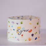 A white buttercream cake with rainbow buttercream dots painted all around the cake, terrazzo style. The rainbow dots resemble paint splotches onna canvas. In the middle of the cake, there is a name "Kyra" written on edible icing sheet.
