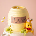 A pale yellow buttercream cake that has been carved into the shape of a honey pot. The cake has an edible label in brown that says "Hunny" in all caps/ There are little fondant bees flying around the cake.