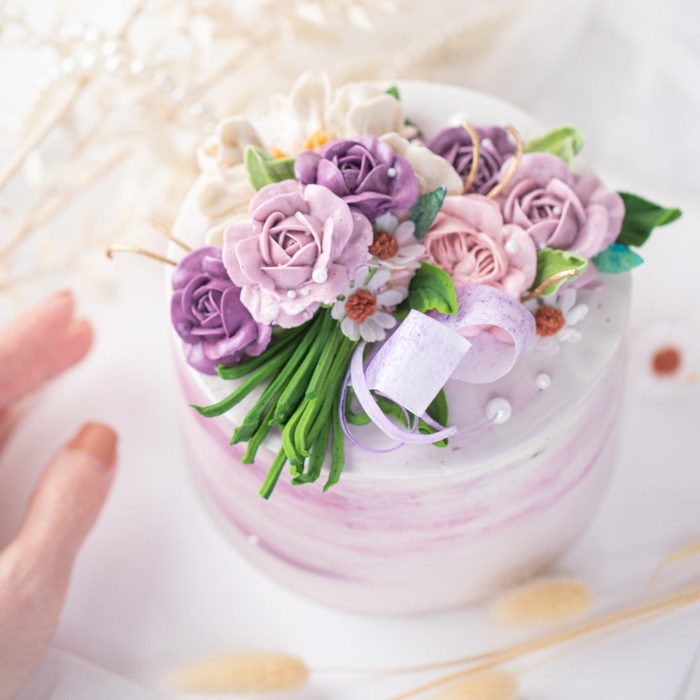 A white buttercream cake with streaks of purple buttercream, and various shades of purple buttercream roses on top of the cake. The buttercream roses have green stems, and resemble a bouquet of flowers. The cake has some edible sugar pearls scattered on top.