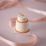 A mini 2 tier white wedding cake. The cake has orange scalloped piping for a contrast, and a mini edible wafer flower on top in orange. The cake is small but beautiful, and incredibly intricate.