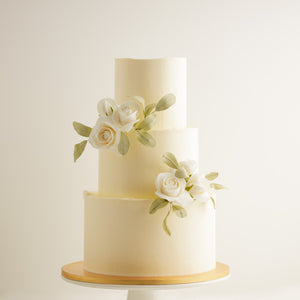 
                  
                    Load image into Gallery viewer, A tall white buttercream cake with three tiers. On the top and bottom of the middle tier, there are some white edible sugar roses that have pale green sugar leaves. The roses look incredibly realistic.
                  
                
