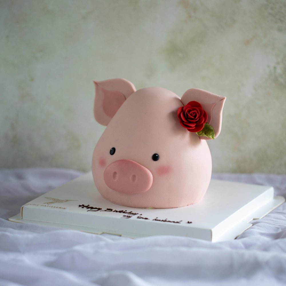 A cake that has been carved and decorated to look like a pig. The cake is covered in light pink fondant, and has adorably cute pig features, including a little fondant nose, small black eyes, and a pink blush. The cake has two fondant pig ears sticking out, and in one of the ears there is a single red fondant rose.