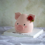 A cake that has been carved and decorated to look like a pig. The cake is covered in light pink fondant, and has adorably cute pig features, including a little fondant nose, small black eyes, and a pink blush. The cake has two fondant pig ears sticking out, and in one of the ears there is a single red fondant rose.