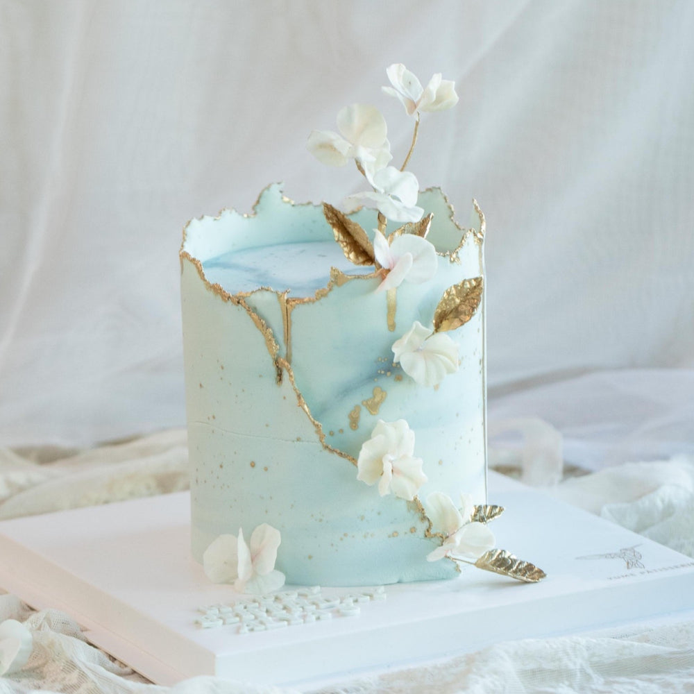 A cake with light blue buttercream carefully wrapped around the side. The buttercream has specks of gold on it, resembling an egg shell. It also has gold luster dust painted on the edges that make the cake pop, along with some edible wafer flowers and gold leaves delicately placed around the cake.