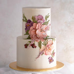 A two tier cake with a white buttercream base, and pink and purple buttercream palette knife flowers on the side of the cake. The flowers are large and detailed, and resemble an oil painting.
