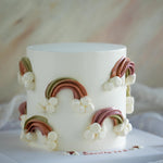 A cake with a smooth white buttercream base that has muted buttercream rainbows in pale red, green and orange piped all around the side. The end of each rainbow has white buttercream clouds piped on.
