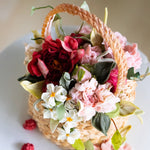 A cake that has been decorated to look like a flower basket. The outside has light brown buttercream that has been piped to resemble a woven basket, and it has assorted buttercream flowers in red, pink and white spilling out of it. It is a realistic flower basket cake.