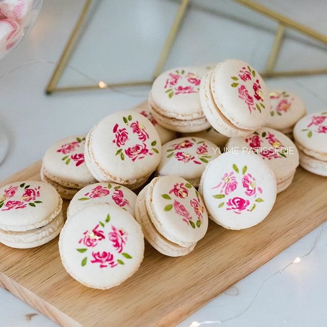 White macarons with hand painted pink flowers in many different designs on them. The macarons also have some gold lead flakes painted on them. 